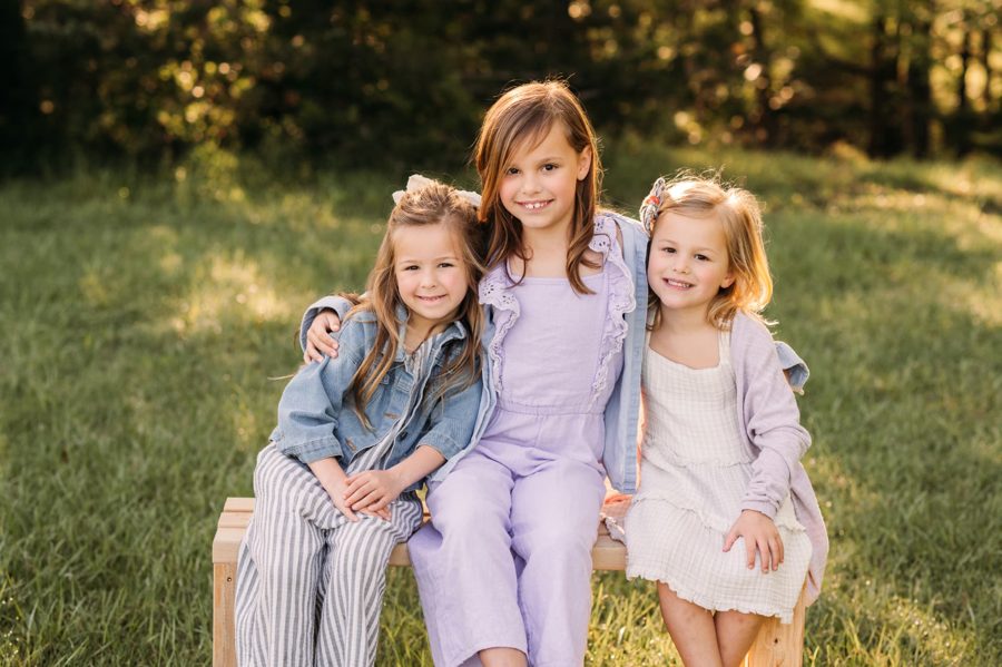 Three sisters sitting together on a wooden bench at golden hour — family photography in the Kansas City area