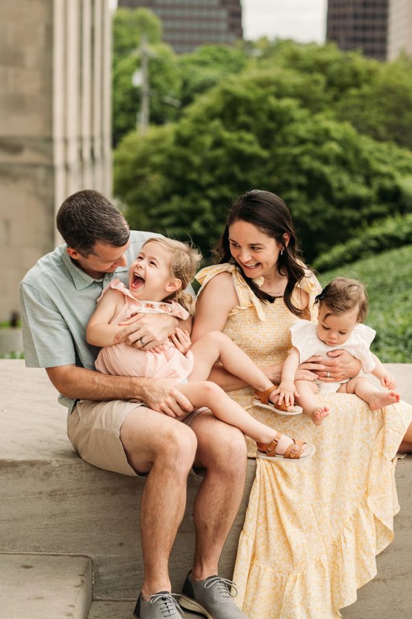 Parents laughing with their two little girls on a stone ledge downtown — family photography in the Kansas City area