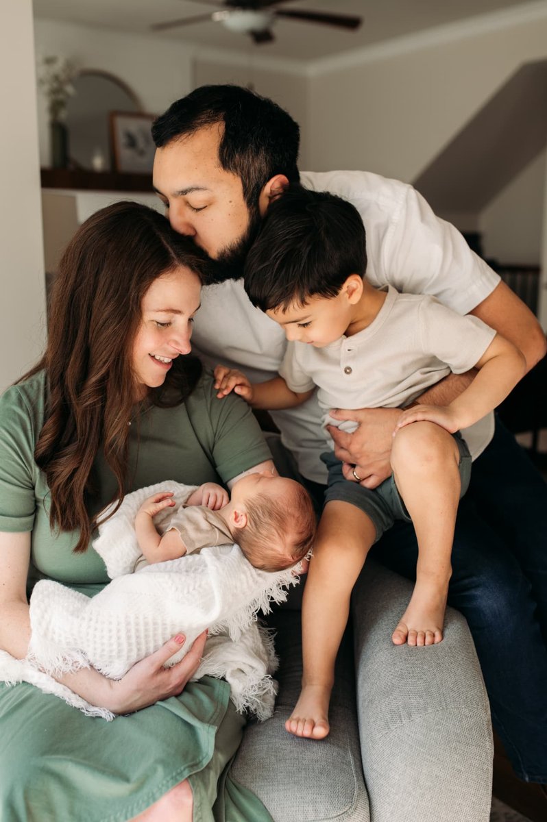 Dad kissing mom while big brother leans in to see the new baby at home — newborn photography in the Kansas City area