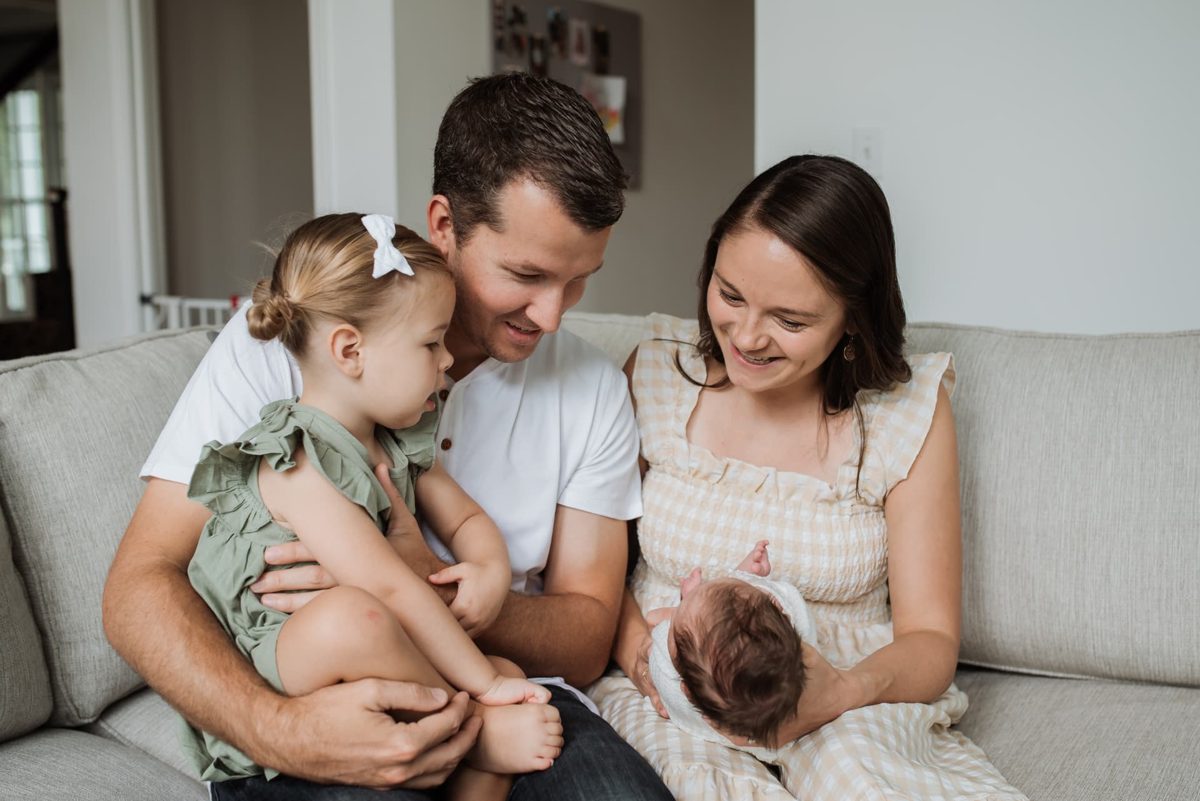 Family of four laughing together on the couch with their newborn daughter — newborn photography in the Kansas City area
