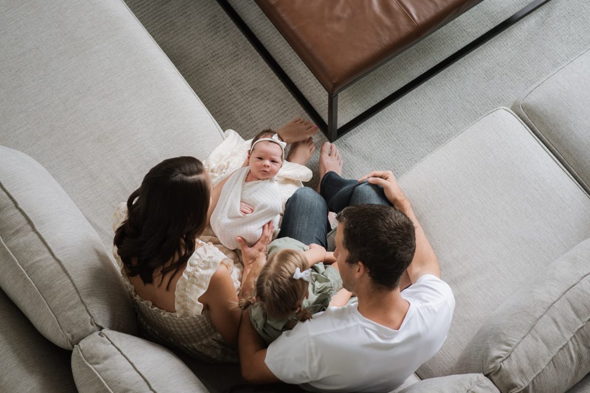 Overhead photo of parents and big sister gathered around their swaddled newborn on the sofa — newborn photography in the Kansas City area