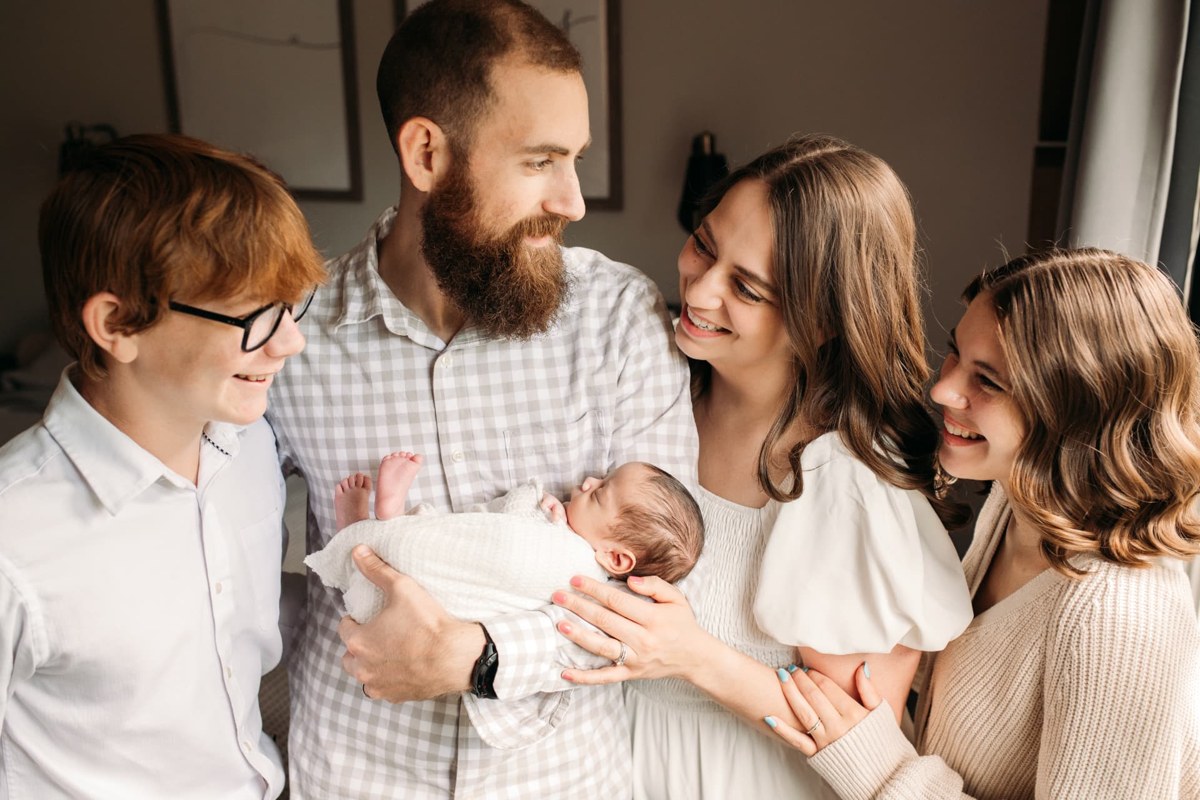 Newborn snuggled in dad's arms while his big siblings and mom smile down — newborn photography in the Kansas City area