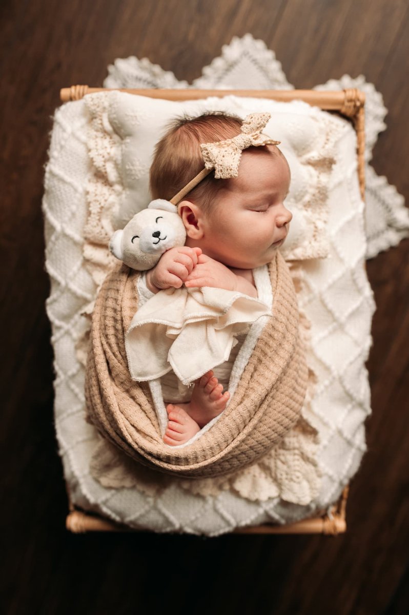 Newborn girl in a lace bow cuddling a little bear in a rattan bed — newborn photography in the Kansas City area