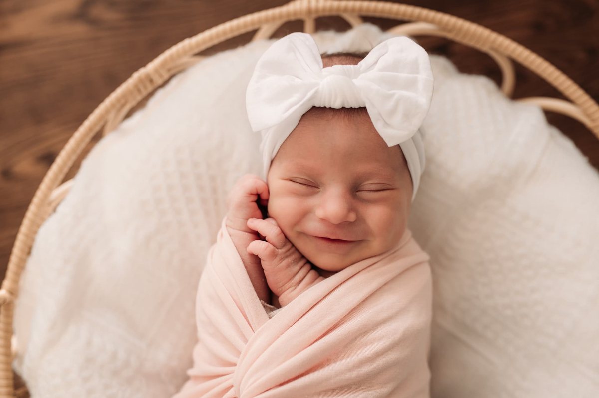 Smiling newborn girl in a pink wrap and white bow in a basket — newborn photography in the Kansas City area