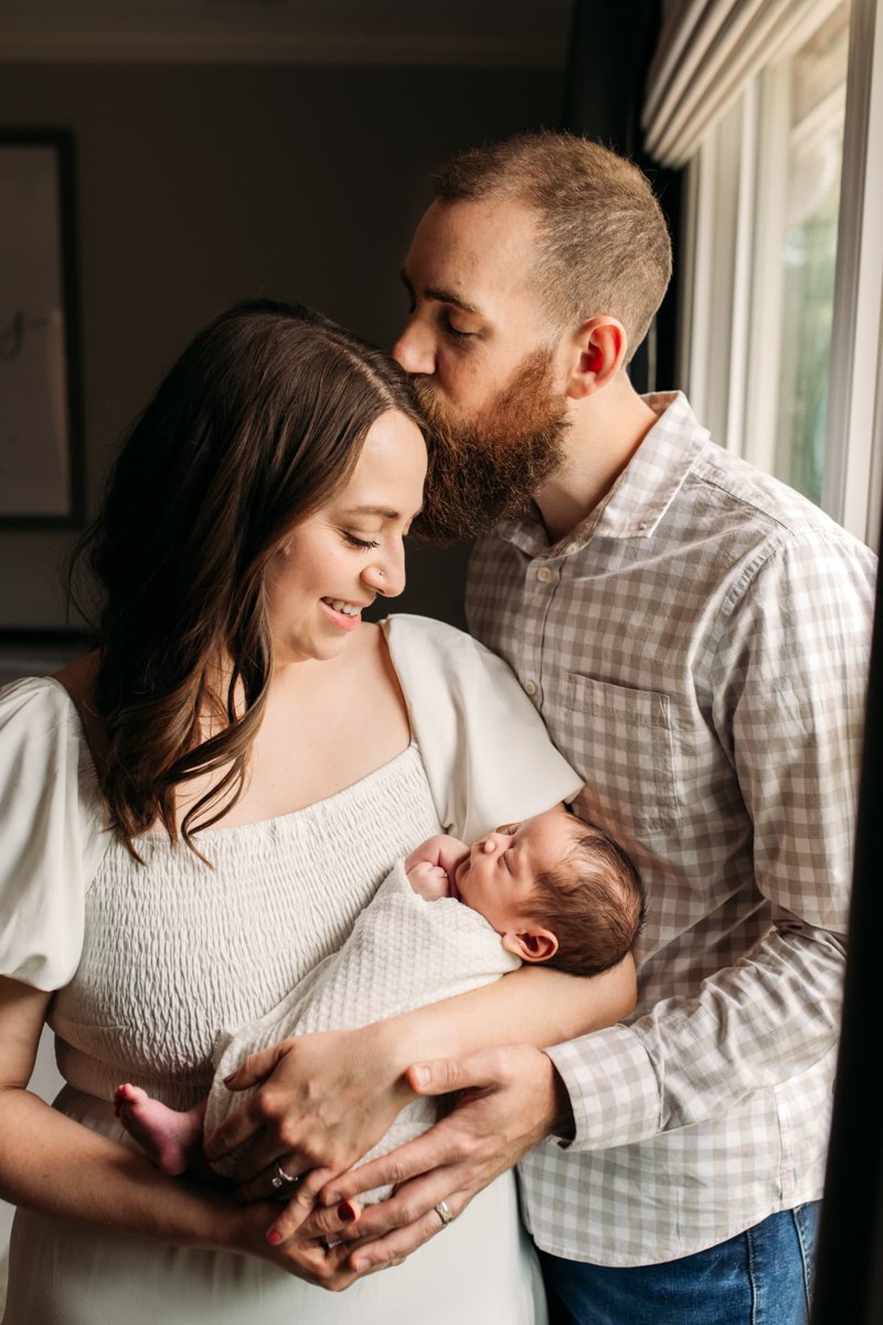 Dad kissing mom's forehead as she holds their swaddled newborn — newborn photography in the Kansas City area
