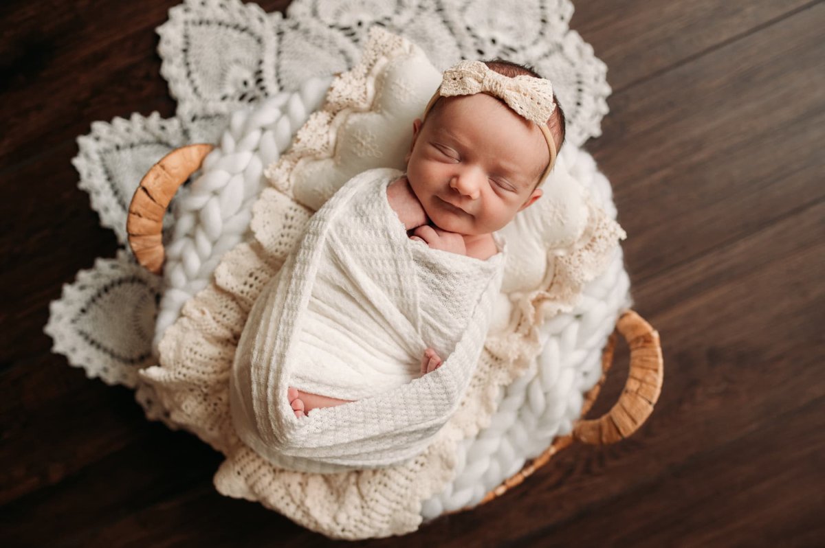 Smiling newborn girl swaddled in cream lace inside a rattan basket — newborn photography in the Kansas City area
