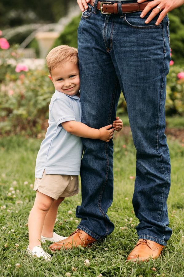 Little boy in a blue polo hugging his dad's leg in the rose garden — family photography in the Kansas City area