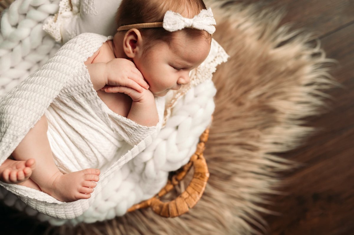 Sleeping newborn girl in a lace bow resting her chin on her hands in a woven basket — newborn photography in the Kansas City area