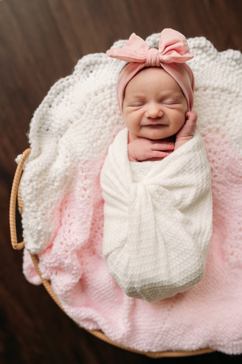 Newborn girl in a pink bow headband grinning in her sleep on a pink knit blanket — newborn photography in the Kansas City area