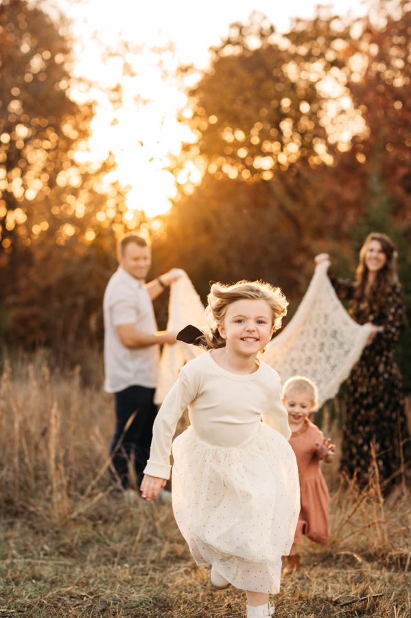 Little girl running toward the camera at sunset with her family behind her — family photography in the Kansas City area