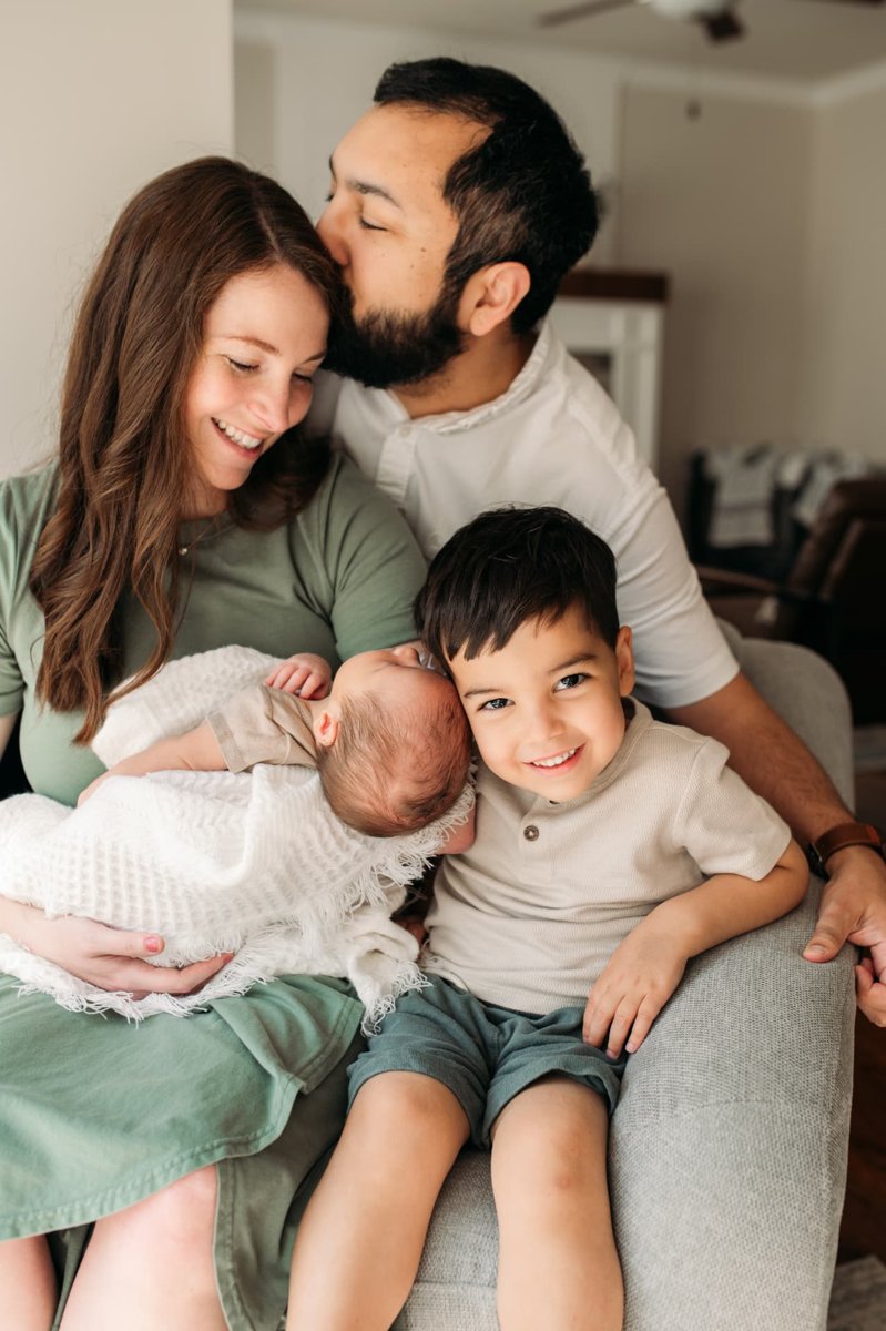 Big brother grinning beside his newborn sibling as dad kisses mom on the couch — newborn photography in the Kansas City area