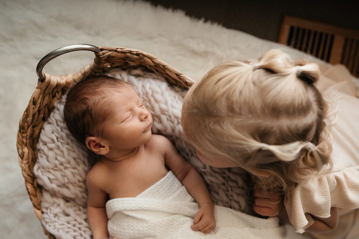 Big sister leaning over her newborn sibling asleep in a basket — newborn photography in the Kansas City area