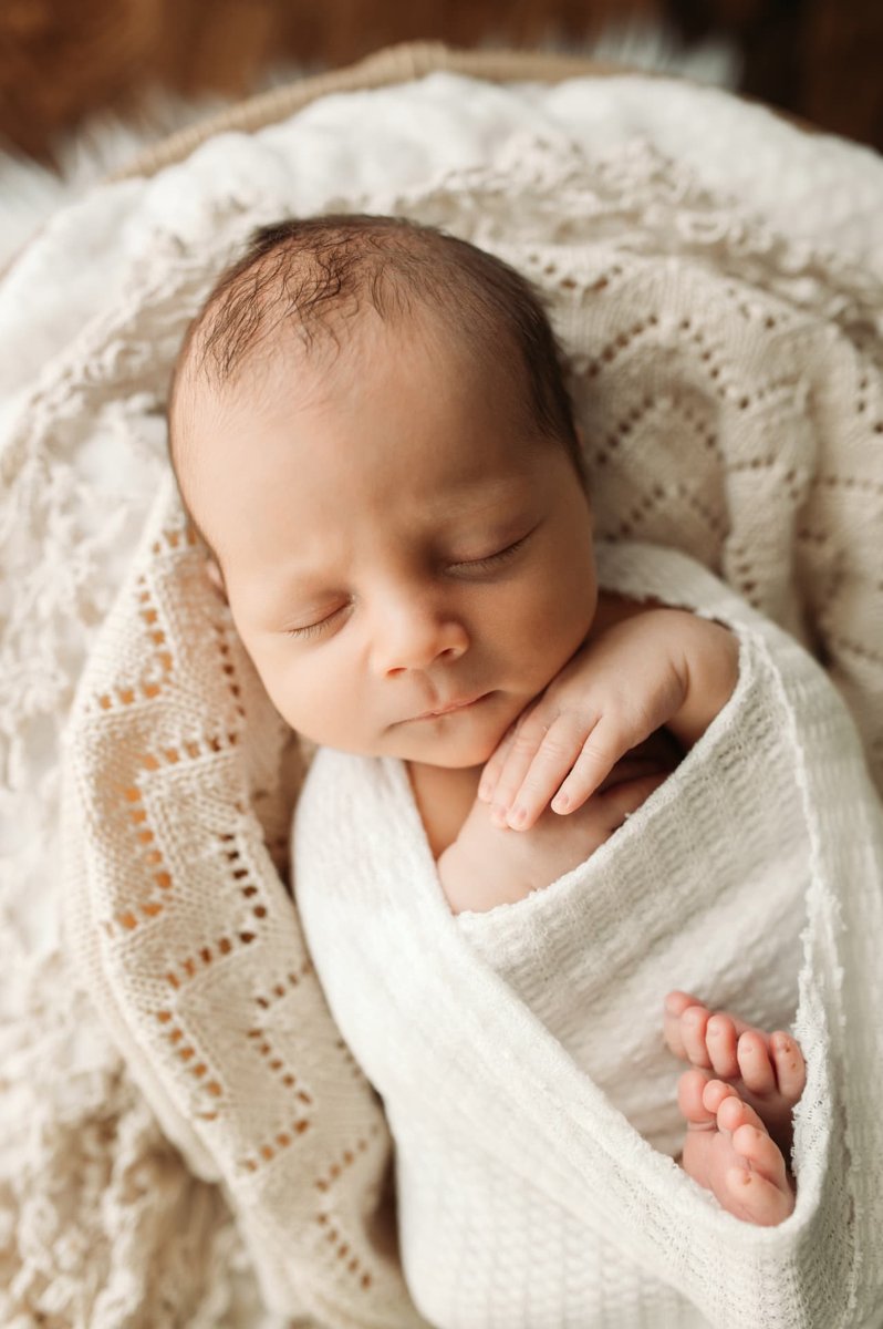 Sleeping newborn swaddled in white lace with tiny toes peeking out in a basket — newborn photography in the Kansas City area