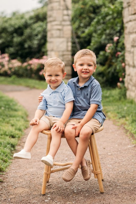 Two little brothers in blue polos sitting together on a rattan stool in the rose garden — family photography in the Kansas City area