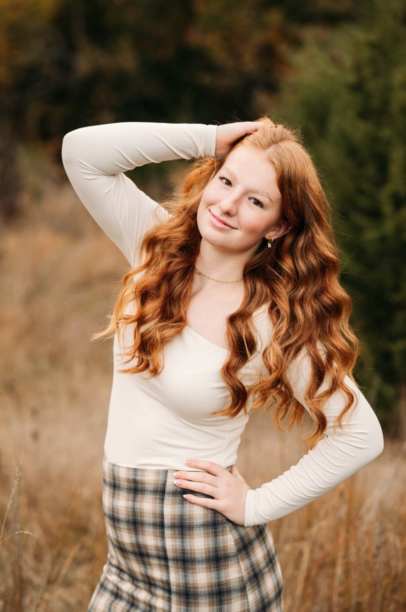 Red-haired senior girl in a cream top and plaid skirt in an autumn field — seniors photography in the Kansas City area