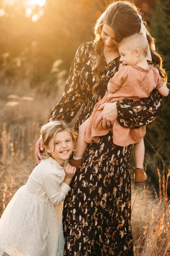 Mother holding her toddler while her daughter hugs her in golden autumn light — family photography in the Kansas City area