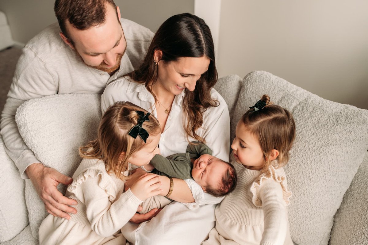 Family of five snuggled on the couch as two big sisters kiss their newborn brother — newborn photography in the Kansas City area