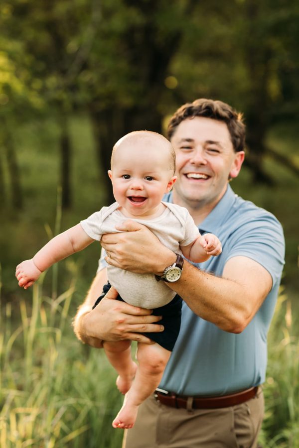 Dad laughing while lifting his baby boy in a sunlit summer field — family photography in the Kansas City area