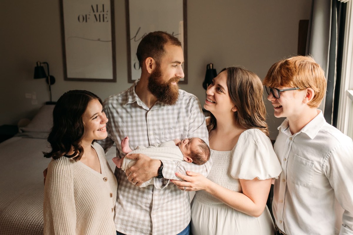 Family of five gathered around their swaddled newborn in a sunlit bedroom — newborn photography in the Kansas City area