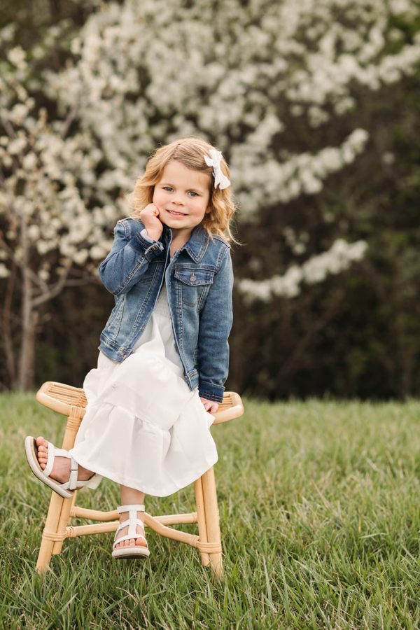 Little girl in a denim jacket sitting on a rattan stool beneath blooming trees — family photography in the Kansas City area