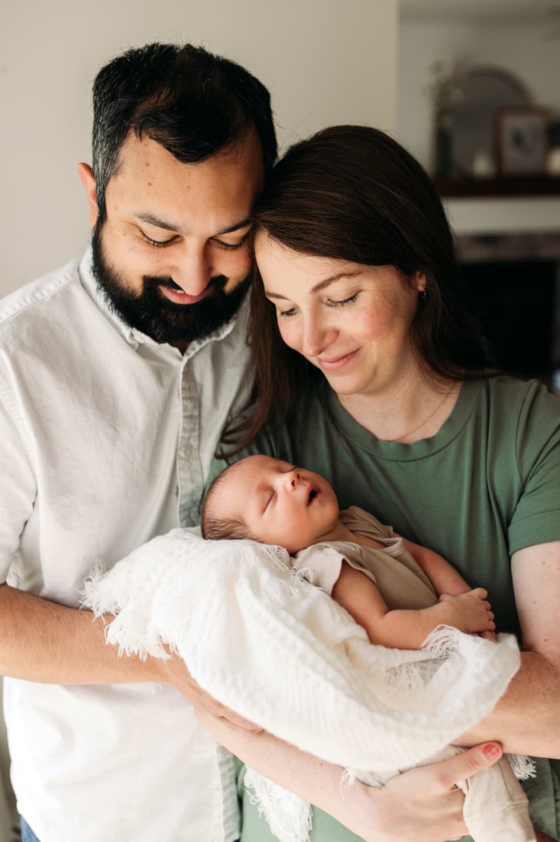Parents cheek to cheek gazing down at their sleeping newborn in a white blanket — newborn photography in the Kansas City area