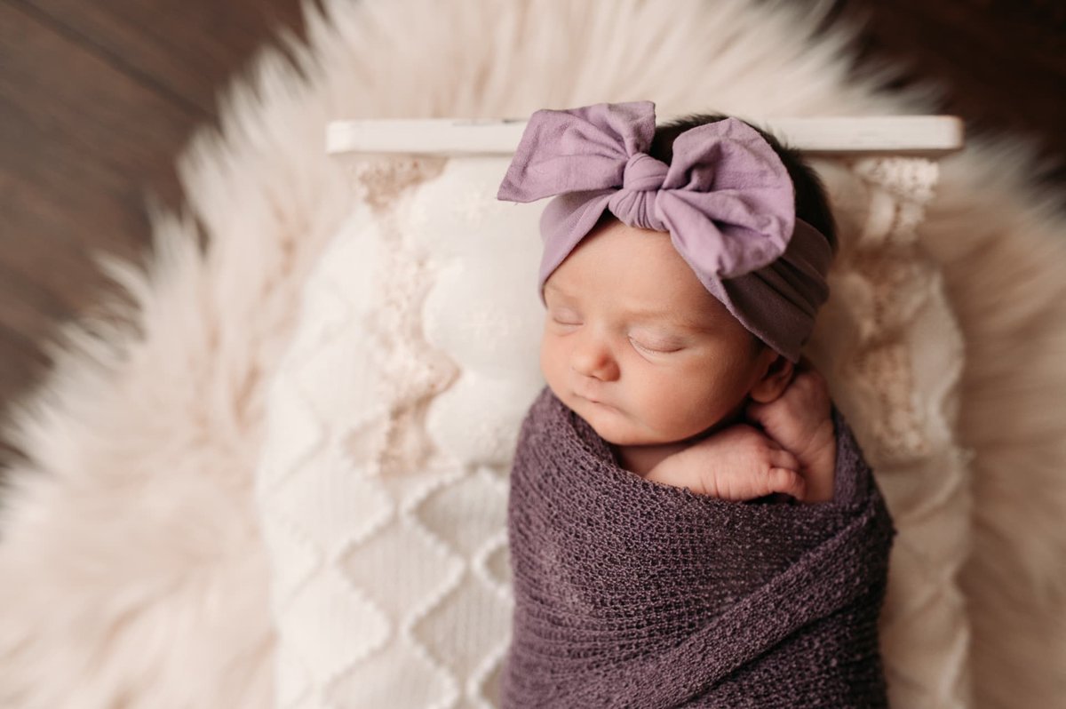Newborn girl in a lavender bow sleeping in a purple wrap on a little white bed — newborn photography in the Kansas City area