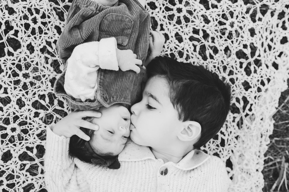 Black and white overhead photo of a big brother kissing his newborn sibling on a lace blanket — newborn photography in the Kansas City area