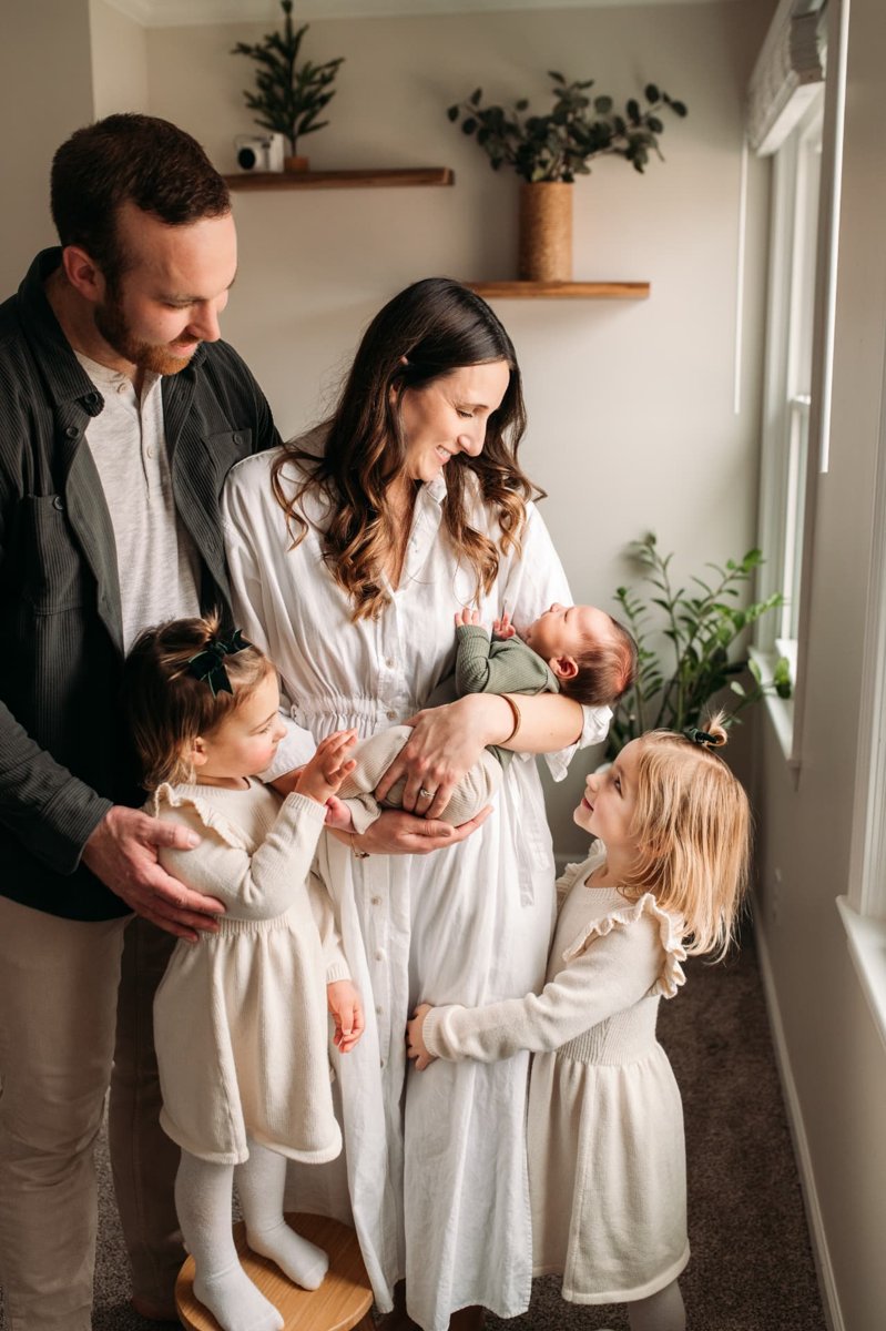 Parents and two daughters gathered in window light with their newborn son — newborn photography in the Kansas City area
