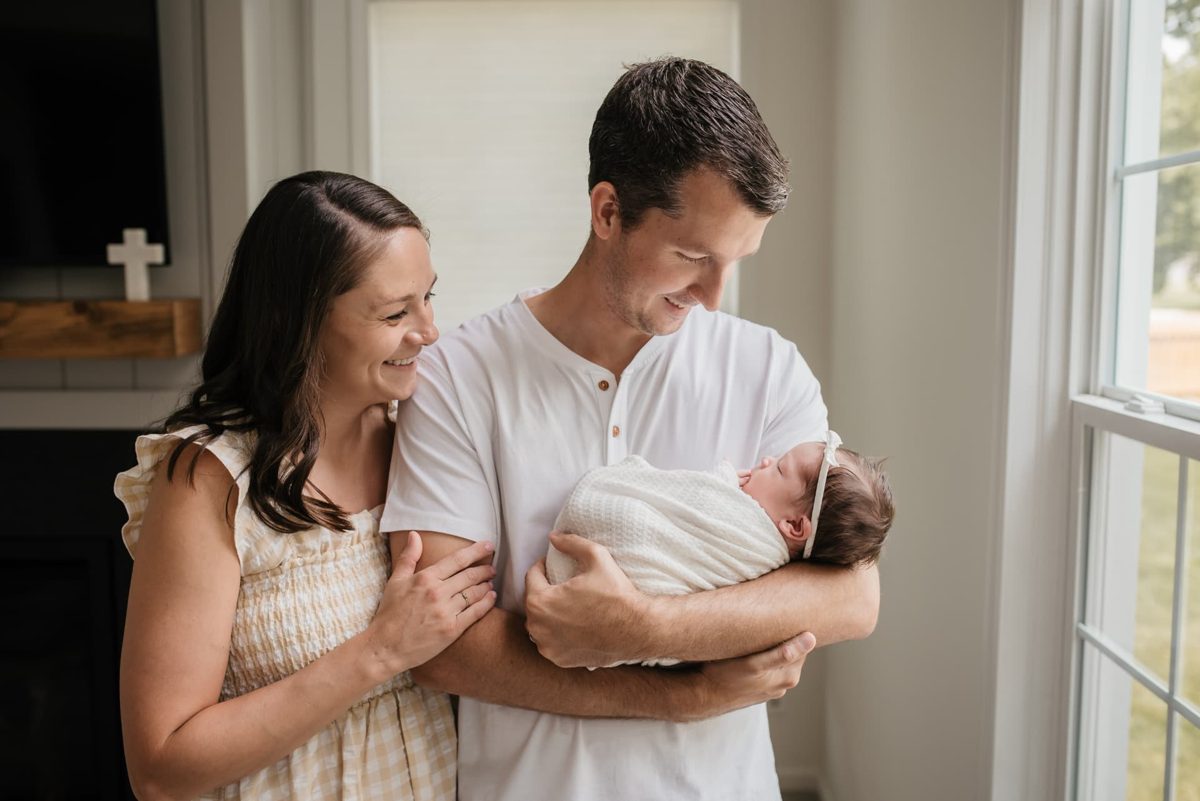 Parents standing by a window holding their swaddled newborn daughter — newborn photography in the Kansas City area