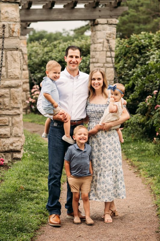 Family of five smiling together under the stone pergola at the rose garden — family photography in the Kansas City area
