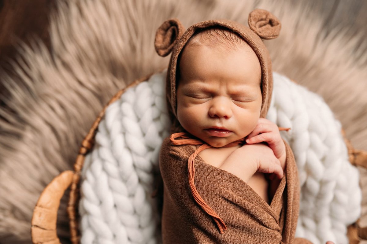 Newborn in a bear-ear bonnet and brown wrap sleeping on a woven basket — newborn photography in the Kansas City area