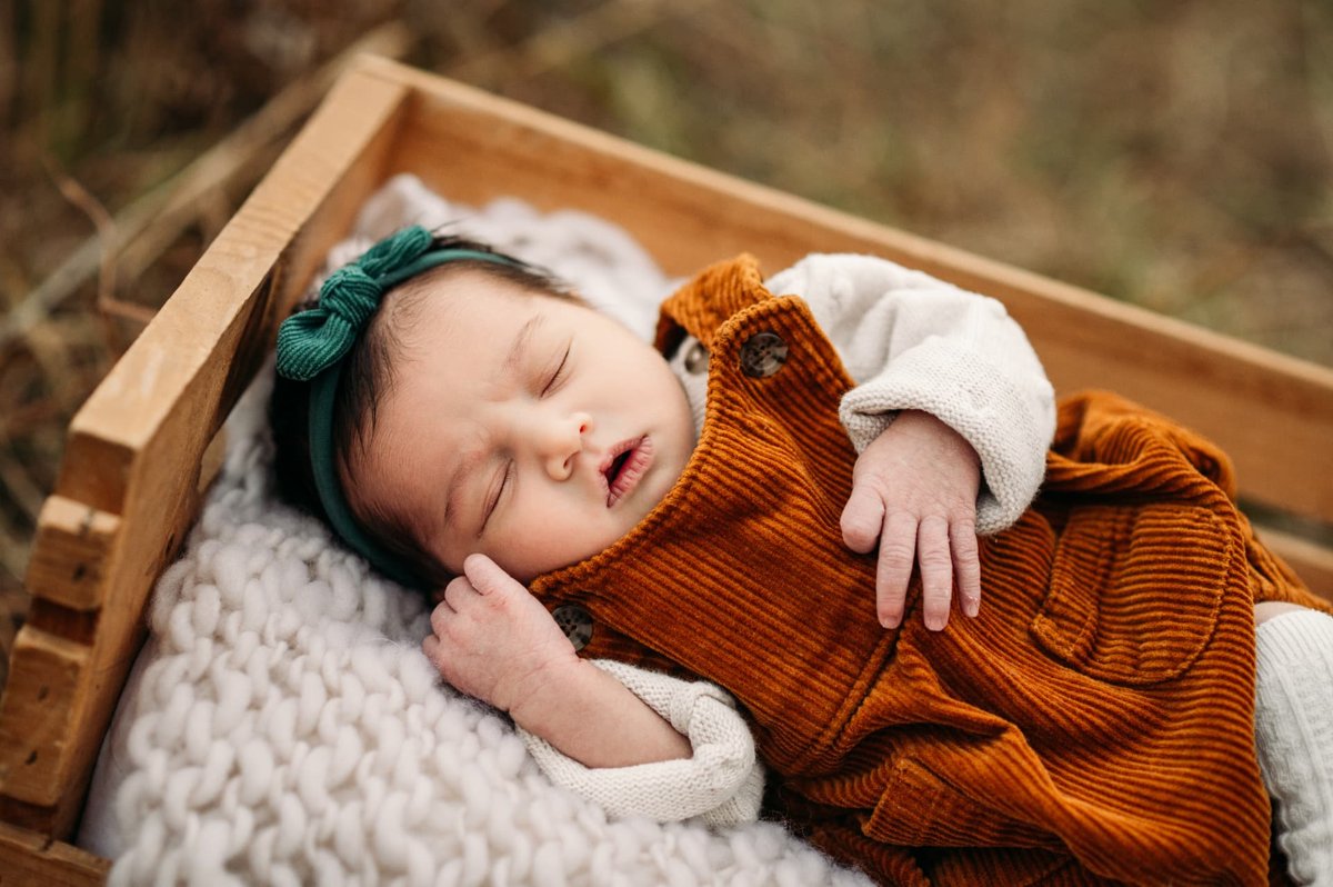 Sleeping newborn girl in rust corduroy overalls in a wooden crate outdoors — newborn photography in the Kansas City area