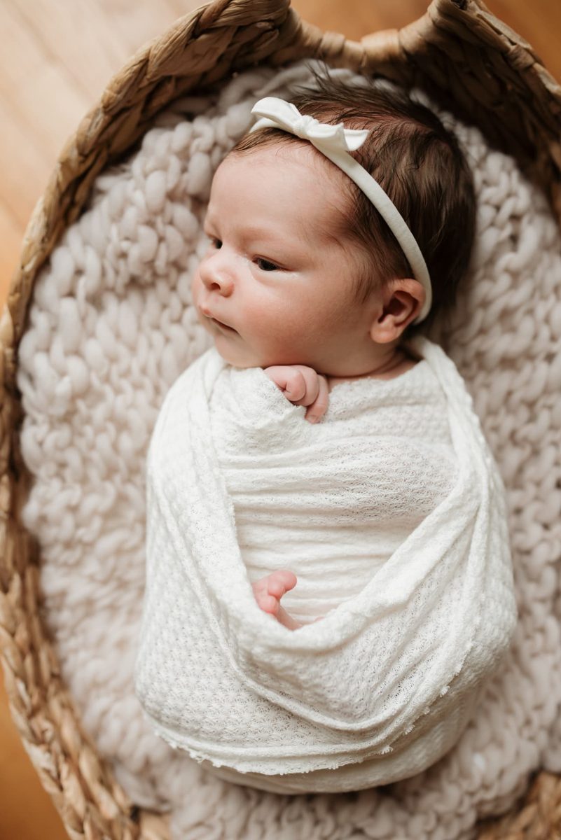 Newborn girl in a bow headband swaddled in a woven basket — newborn photography in the Kansas City area