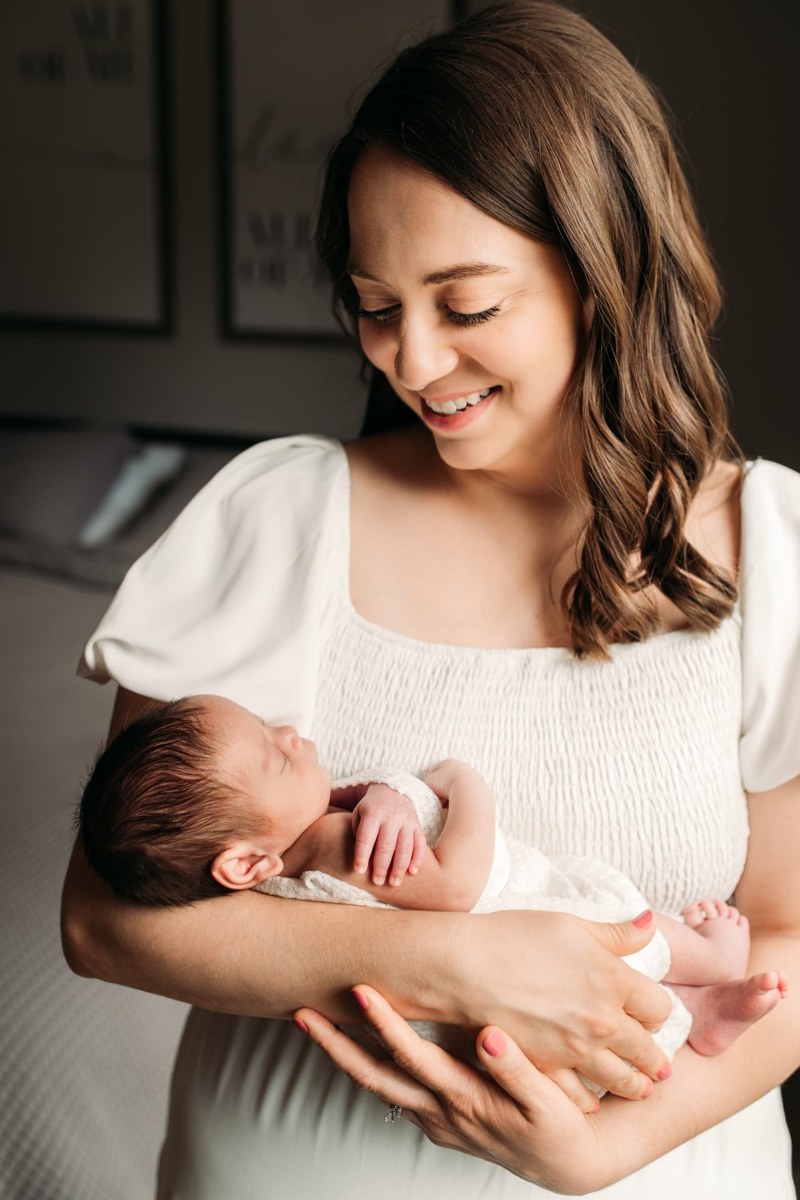 Mom smiling down at her sleeping newborn in soft window light — newborn photography in the Kansas City area