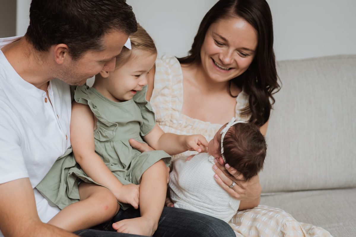 Big sister reaching for her newborn sister while mom and dad smile — newborn photography in the Kansas City area