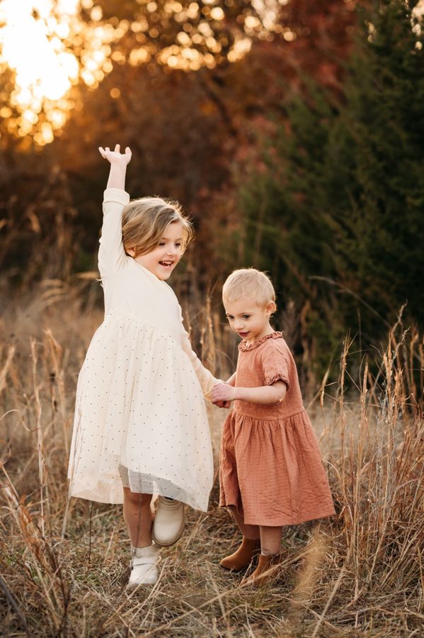 Sisters holding hands and dancing in a golden autumn field — family photography in the Kansas City area
