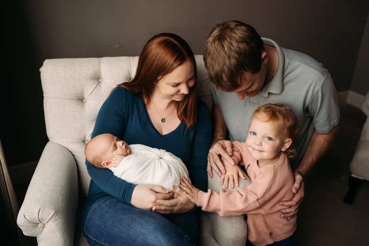 Parents and big sister gathered around their swaddled newborn in a nursery chair — newborn photography in the Kansas City area