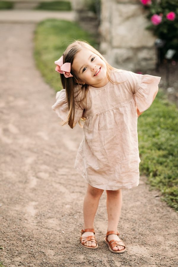 Little girl in a linen dress tilting her head and smiling on a garden path — family photography in the Kansas City area