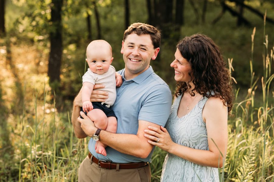 Dad holding his baby boy while mom laughs beside them in tall grasses — family photography in the Kansas City area
