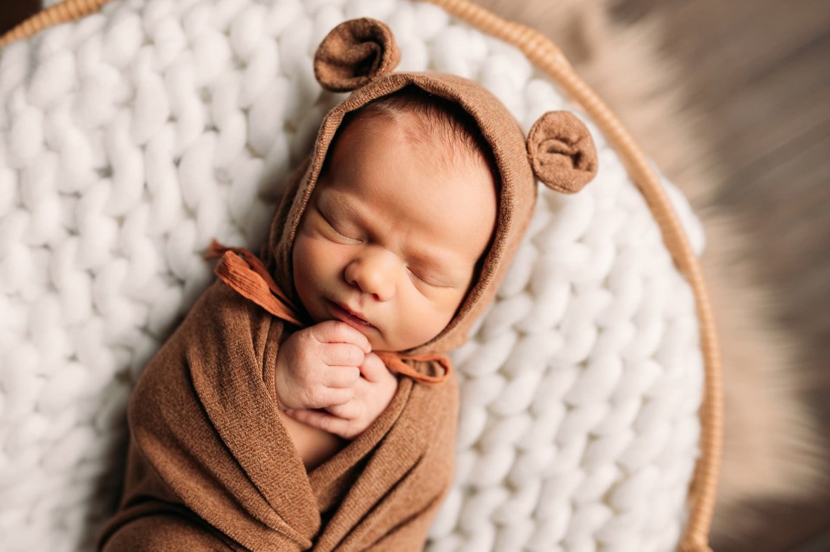 Sleeping newborn in a brown bear bonnet curled in a chunky knit basket — newborn photography in the Kansas City area