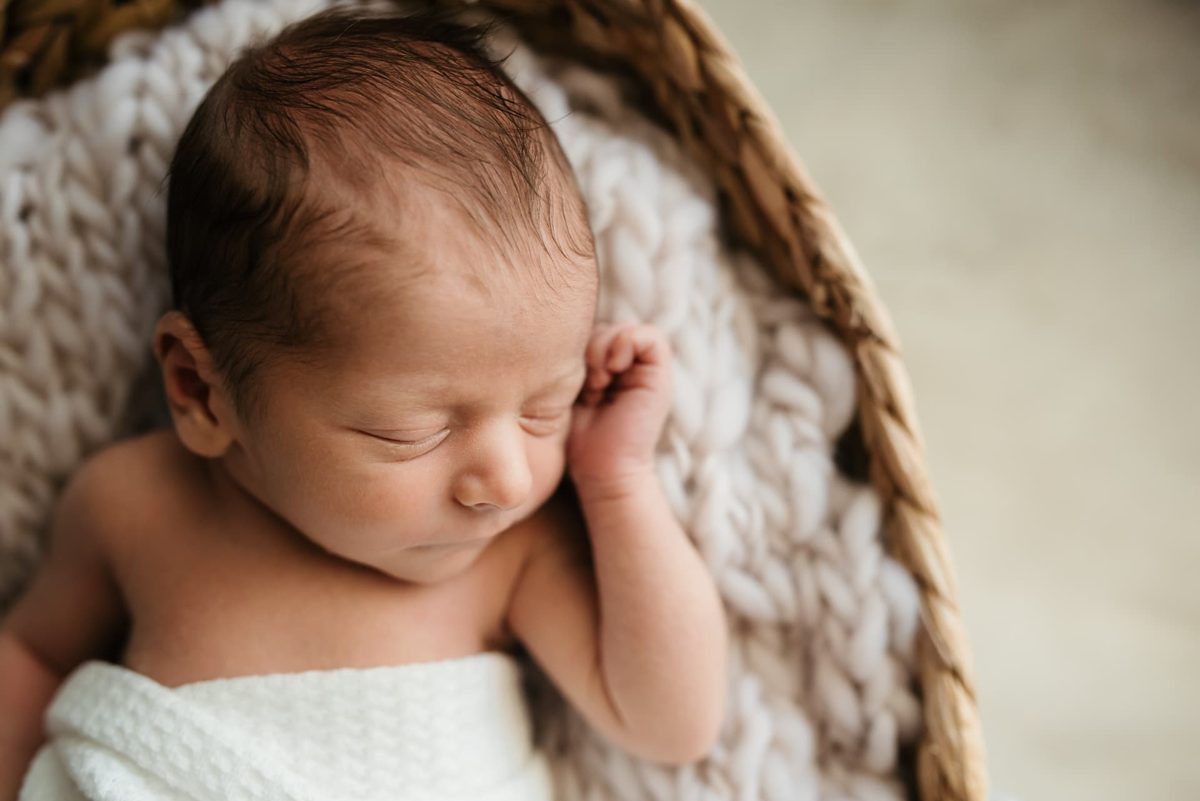 Sleeping newborn resting a hand beside her cheek in a knit-lined basket — newborn photography in the Kansas City area