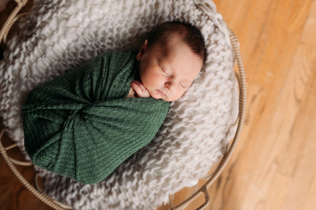 Newborn wrapped in a green knit swaddle asleep in a basket — newborn photography in the Kansas City area