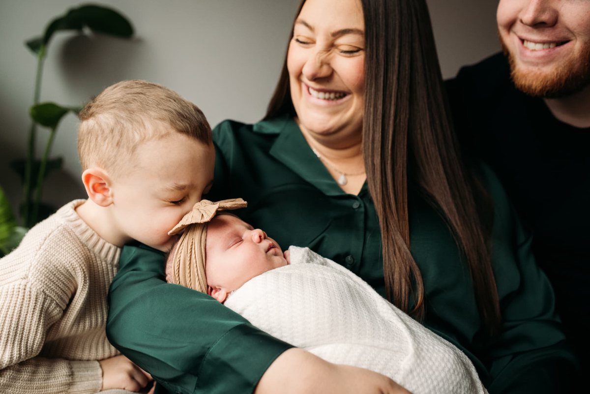 Big brother kissing his swaddled newborn sister as parents smile