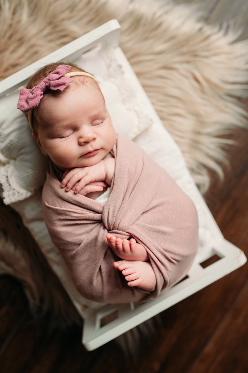 Newborn girl in a mauve wrap and bow sleeping on a tiny white bed — newborn photography in the Kansas City area
